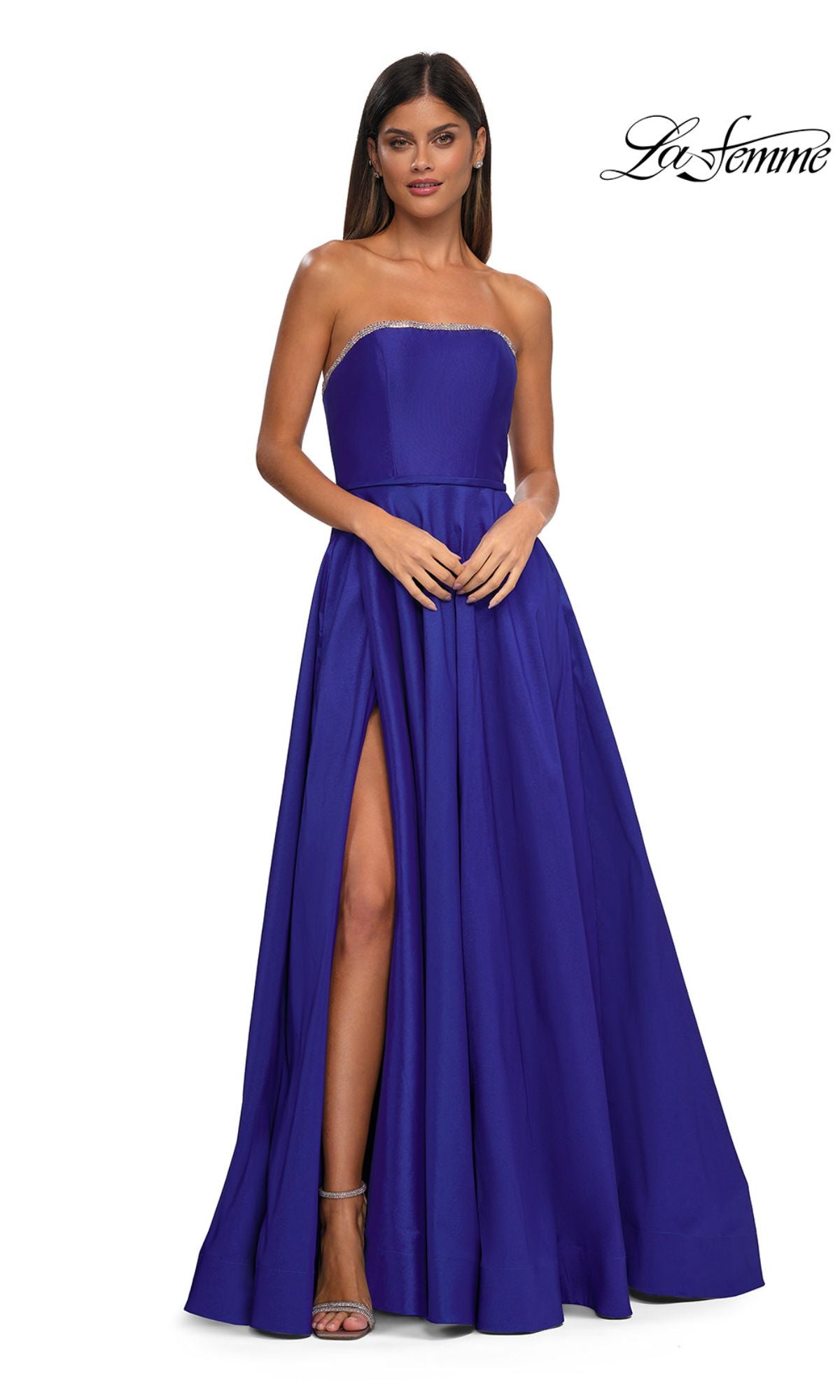 A woman wearing the Strapless Long A-Line Prom Dress La Femme 33063 in royal blue, featuring rhinestone beading and a thigh-high slit, paired with silver high heels, stands against a white background.